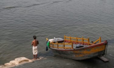 A man stands by a riverbank near a vibrant painted boat in Mathura, India.