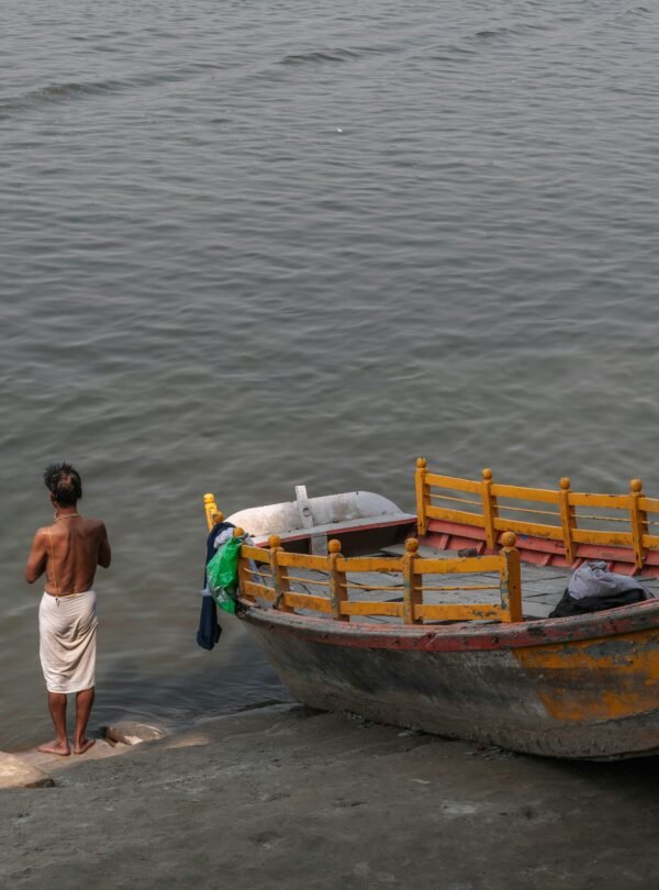 A man stands by a riverbank near a vibrant painted boat in Mathura, India.